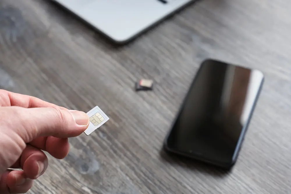 Main masculine tenant une carte SIM pour un smartphone, sur fond de table en bois, téléphone et ordinateur portable. Remplacement du numéro Abonné, mise au point sélective.